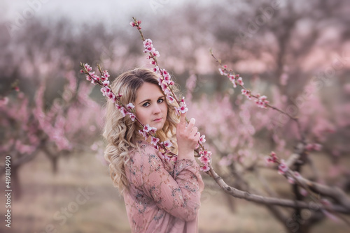 Tender young woman with beautiful curly blonde hair, with clean face, natural makeup, stands in blooming gardens, holds twig with pink inflorescences. Mysterious, pensive, sensitive girl. Close up