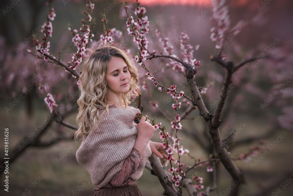 Young beautiful curly blonde woman in brown pleated skirt pink blouse covered shoulders with knitted scarf, stands in blooming peach sunset gardens. Girl enjoying walk outside the city, natural beauty Young beautiful curly blonde woman in brown pleated skirt pink blouse covered shoulders with knitted scarf, stands in blooming peach sunset gardens. Girl enjoying walk outside the city, natural beauty