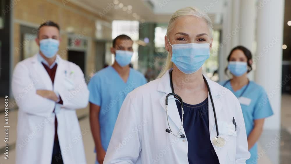 Satisfied senior doctor wearing face mask with team in background Satisfied senior doctor wearing face mask with team in background