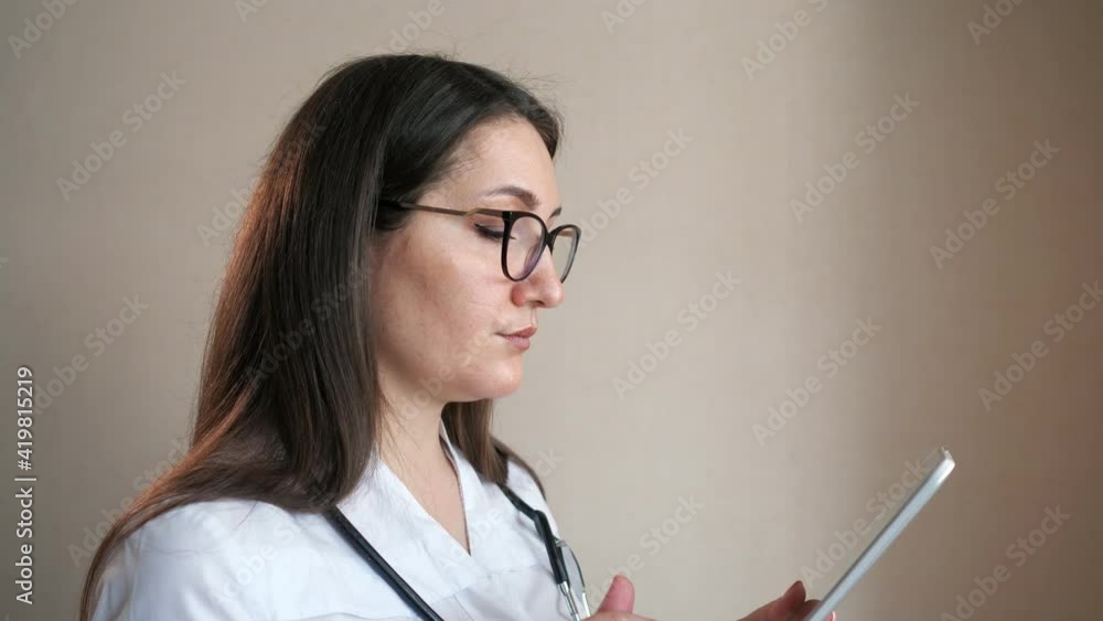 Concentrated young woman doctor with glasses and stethoscope uses contemporary tablet standing near beige wall in clinic office close view Concentrated young woman doctor with glasses and stethoscope uses contemporary tablet standing near beige wall in clinic office close view