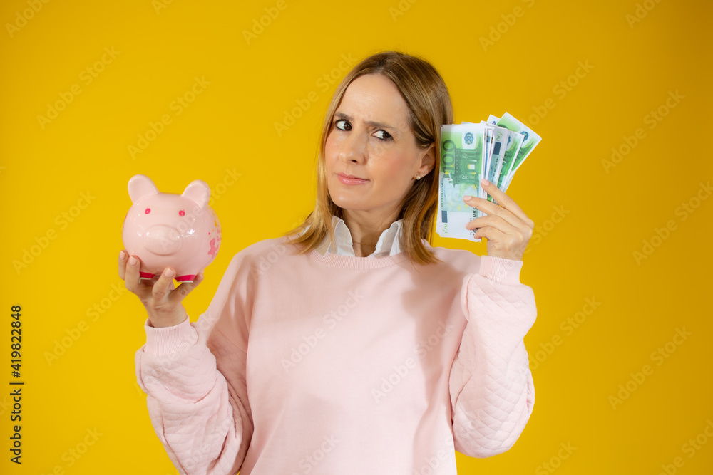 Portrait of a pensive young woman holding piggy bank and bunch of money banknotes isolated over yellow background Portrait of a pensive young woman holding piggy bank and bunch of money banknotes isolated over yellow background