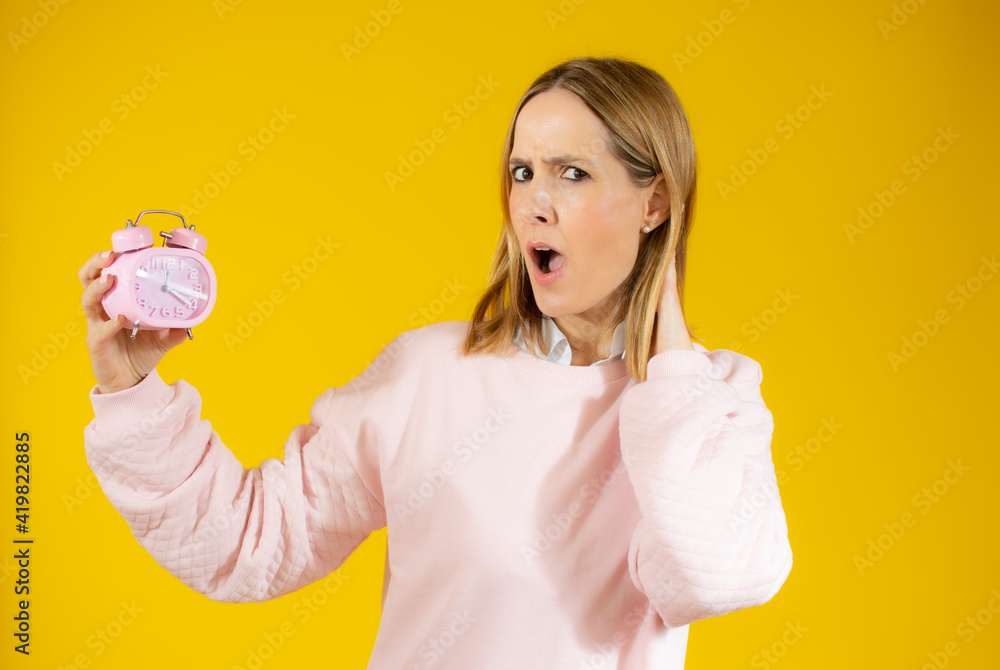 Young woman surprised holding alarm clock sanding over yellow background Young woman surprised holding alarm clock sanding over yellow background