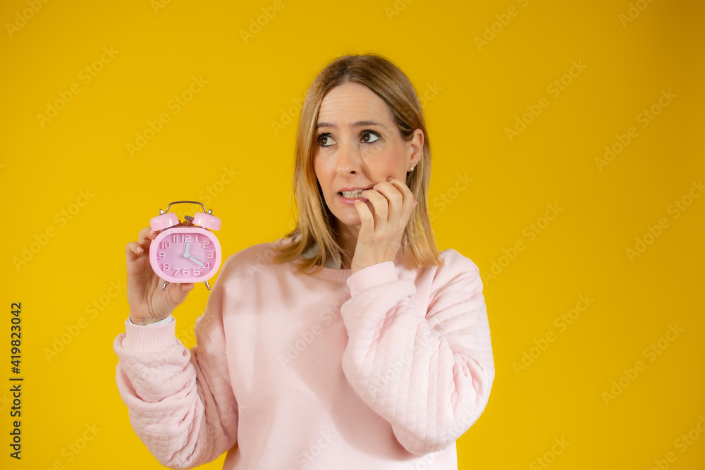 Beautiful young woman holding an alarm clock with worried gesture over yellow background Beautiful young woman holding an alarm clock with worried gesture over yellow background
