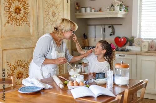 Φωτογραφία Grandma in white apron with cute curly granddaughter spend wonderful time in the kitchen, smiling and there are happy