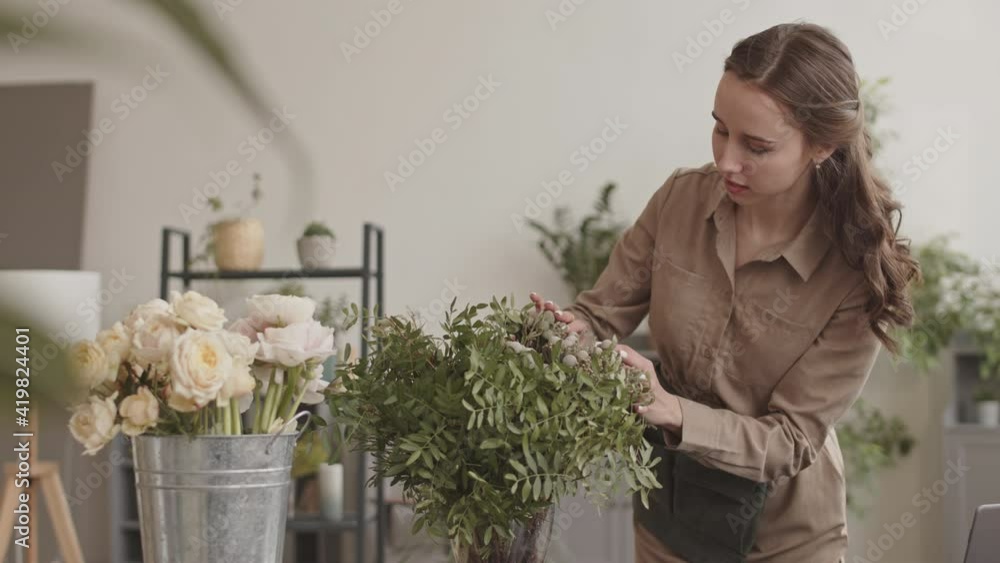 Medium long of young Caucasian woman wearing light-brown dress, standing behind counter in flower shop, touching up bouquet of decorative greens in vase, smiling Medium long of young Caucasian woman wearing light-brown dress, standing behind counter in flower shop, touching up bouquet of decorative greens in vase, smiling