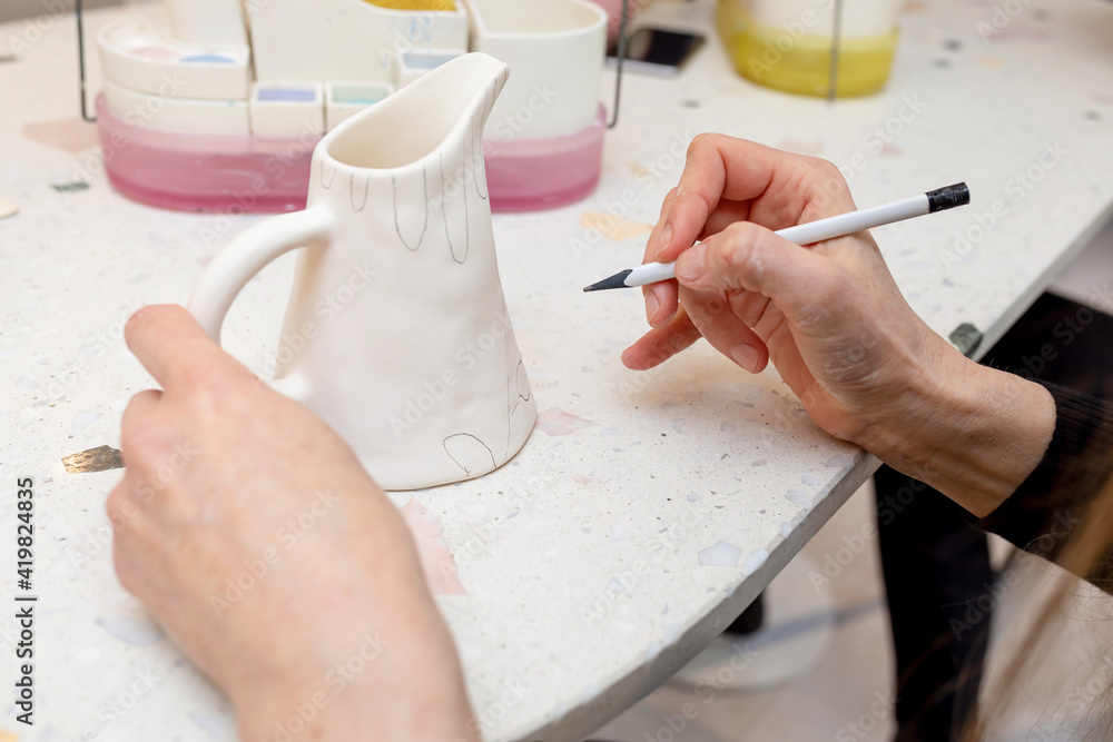 A woman applies a pencil sketch on a white ceramic jug before applying glazed paints. The concept of manual creativity, leisure, hobby