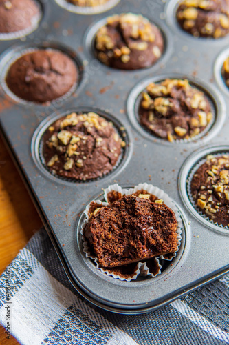 Selective focus on a half eaten healthy gluten free chocolate muffin with walnuts. Homemade, freshly baked goods with cocoa powder and dark chocolate placed in a muffins tray in the background.