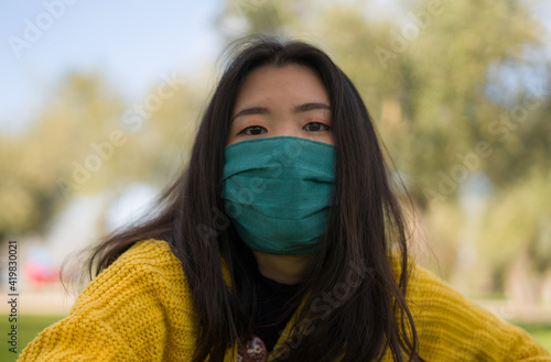 young Asian woman in city park during covid19 - outdoors lifestyle portrait of happy and pretty Korean girl in face mask sitting relaxed on grass during virus time © TheVisualsYouNeed