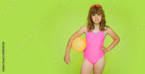 Mockup children's swimsuit. A beautiful little pre-adolescent girl in a pink swimsuit on a green background looks at the camera. Cute baby with swimming goggles and beach volleyball. Place for text