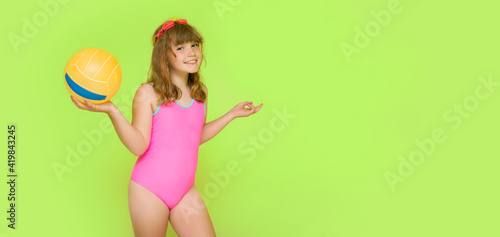 A beautiful child in a pink swimsuit and with a beach volleyball ball in his hand. Plortret of a cute little girl with swimming goggles on a green background. Summer vacation concept. Place for text