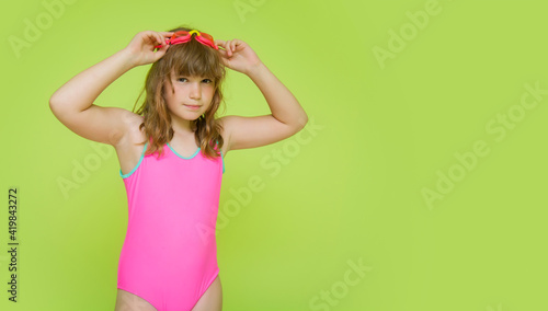 A beautiful pre-teen girl in a pink colorful bathing suit getting ready to swim in the pool. Mockup. Funny child dressed in bright swimwear and glasses on a green background. Place for text