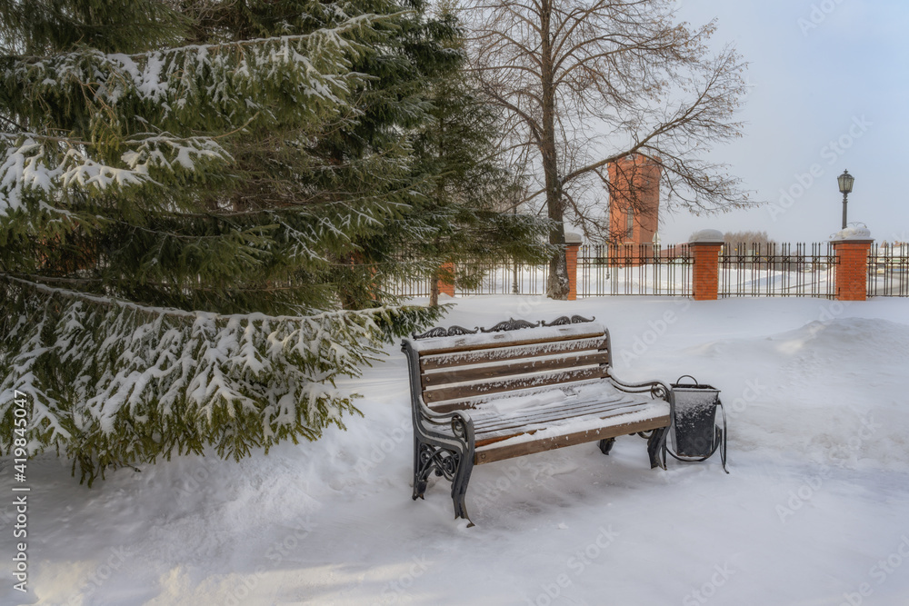 In a winter park with old century-old fir trees. A cozy wooden bench for relaxation and romantic meetings. An old brick water tower rises in the distance. Photo taken in Tobolsk (Siberia, Russia) 