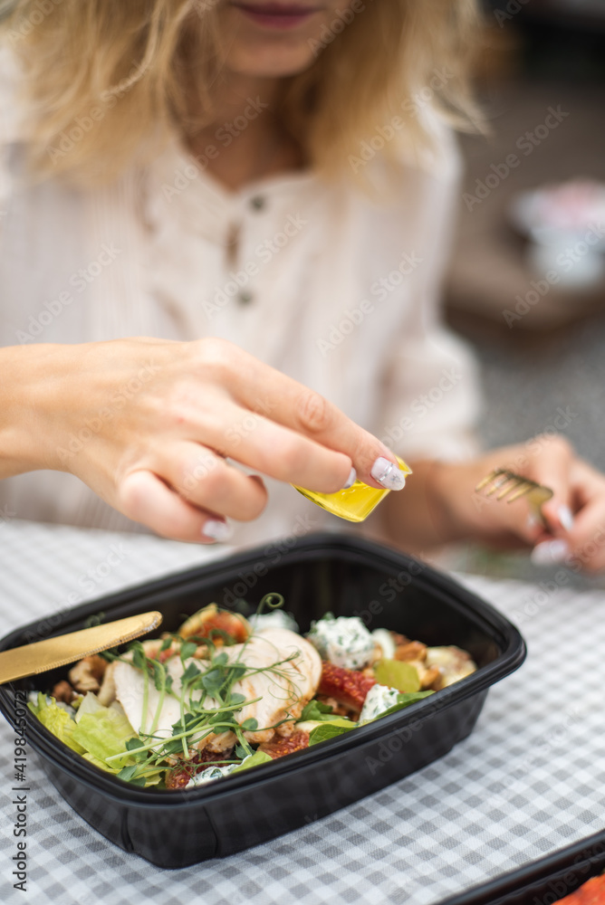 Woman hand pouring flavored olive oil to fresh vegetable salad. Preparation healthy vegetarian food for garden party outdoors. The salad is in a food container. Food delivery.
