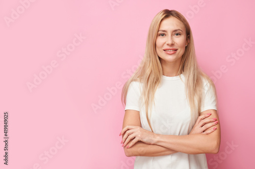 portrait beautiful young blond woman in casual toothy smiling with arms crossed and looking at the camera isolated on pink studio background with copy space