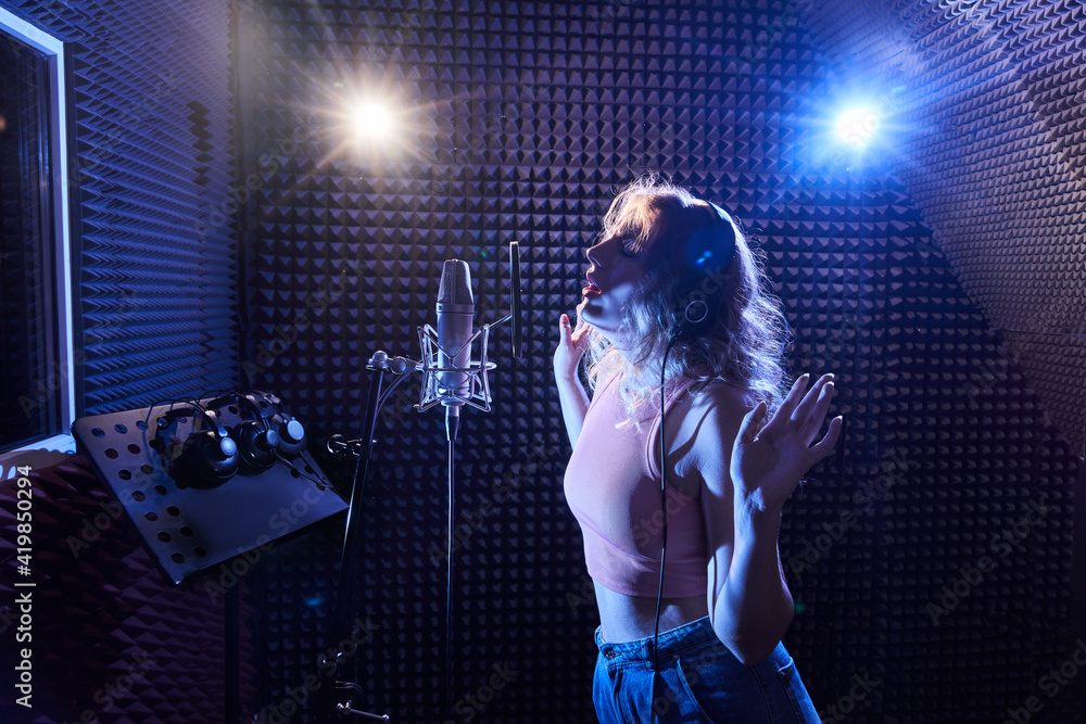Obraz premium Beautiful blonde girl emotionally singing song in recording studio with professional microphone and headphones, creates new track album, vocal artist in pink blue neon light