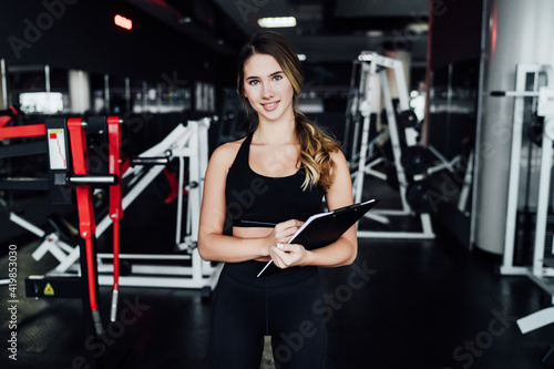 Fitness blonde girl personal fitness trainer with workout plan in hand, smiling and looking at camera, she is in the middle of modern gym
