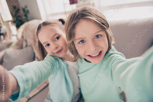 Photo portrait of small sister and brother taking selfie sitting on couch together spending at time at home