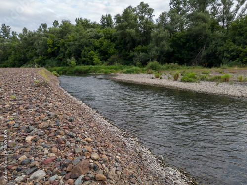 Landscape of river Ukrine, fast water current eroding gravel shore, riverbank overgrown with green bushes and trees during overcast summer day
