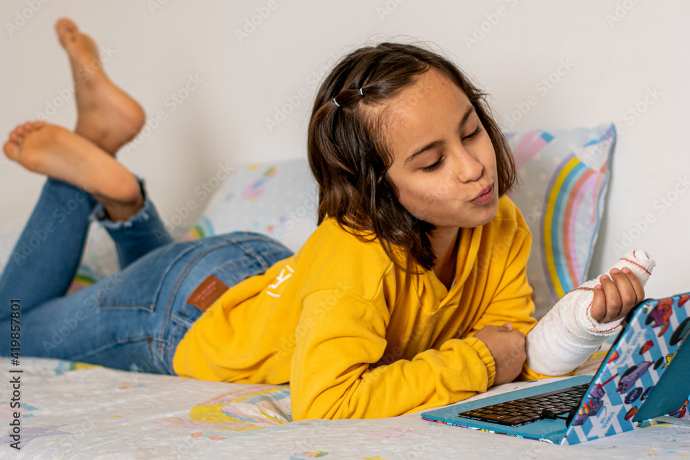 Little Caucasian girl, with a cast in one hand, lying on the bed making a video call with friends with her tablet. Little Caucasian girl, with a cast in one hand, lying on the bed making a video call with friends with her tablet.