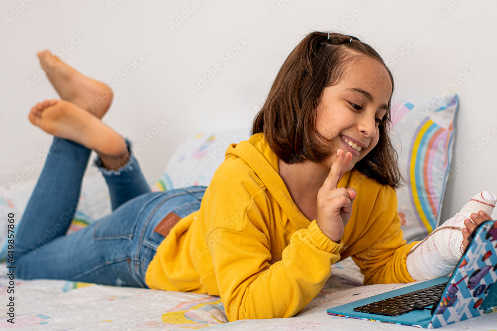 Little Caucasian girl, with a cast in one hand, lying on the bed making a video call with friends with her tablet. Little Caucasian girl, with a cast in one hand, lying on the bed making a video call with friends with her tablet.