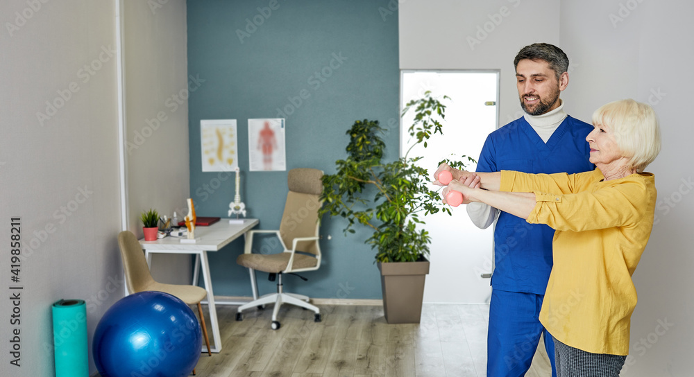 Fototapeta premium Older woman trains with physiotherapist using dumbbells at a medical center