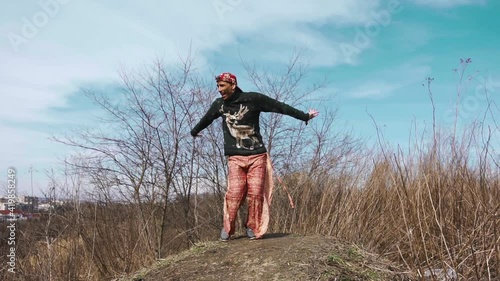 A white man with dark paint on his face and a cap on his head and in strange wide pants runs up and jumps up the hill and tries to dance an Indian dance and waves his hands incomprehensibly photo