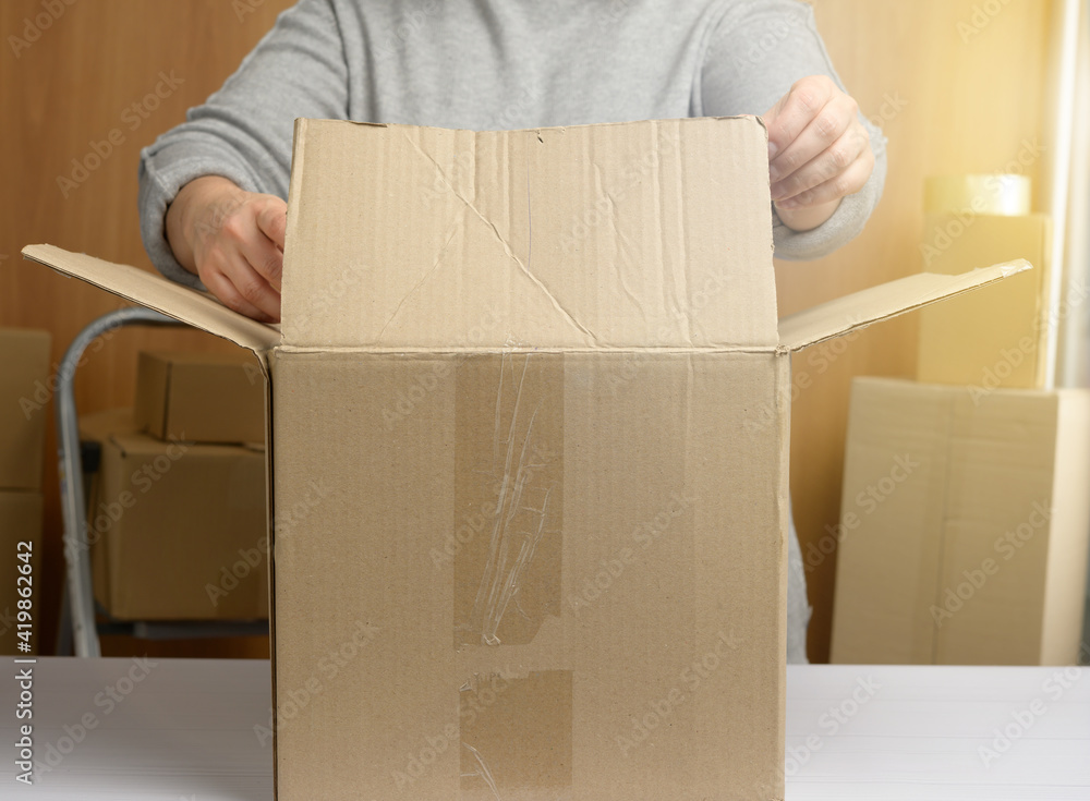 woman in a gray sweater is packing brown cardboard boxes on a white table, behind a stack of boxes woman in a gray sweater is packing brown cardboard boxes on a white table, behind a stack of boxes