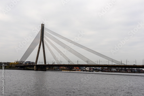 Riga cable-stayed bridge over Daugava river during grey autumn day when water, sky and bridge are depressing grey, but some colourful houses are visible other side of river.