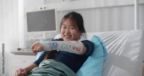 Quadro em tela Smiling asian preteen girl lying in hospital bed with broken arm in colored cast