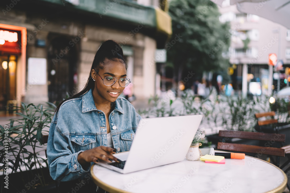 Obraz premium Dark skinned woman in glasses typing on laptop computer share content in blog browsing web pages and information,African American female freelancer doing remote job using wifi internet at cafe terrace