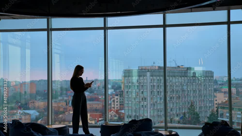 Full-length portrait on a business woman in modern office. Panoramic view on large office windows with cityscape indoors. Beautiful female business worker with a phone. Full-length portrait on a business woman in modern office. Panoramic view on large office windows with cityscape indoors. Beautiful female business worker with a phone.