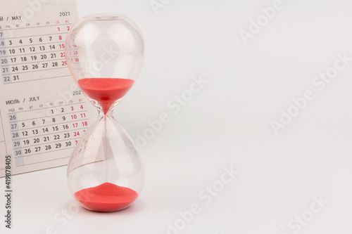 Time passing concept. Crystal hourglass with red sand and calendar on light background