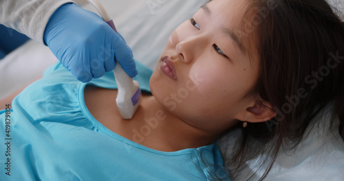 Preteen asian girl lying on bed while getting ultrasound of thyroid from doctor at hospital