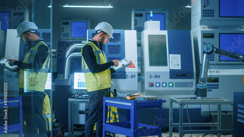 Modern Factory: Professional Engineer Wearing Safety Vest and Hardhat, Uses Industrial Digital Tablet to Program and Manipulate Robot Arm, Optimizing Production Line. Workshop with CNC Machinery