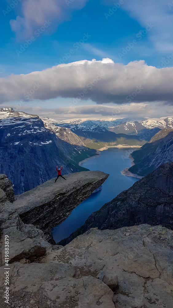 A girl wearing pink jacket jumps at the hanging rock formation, Trolltunga with a view on Ringedalsvatnet lake, Norway. Slopes of the mountains are partially covered with snow. Freedom and happiness