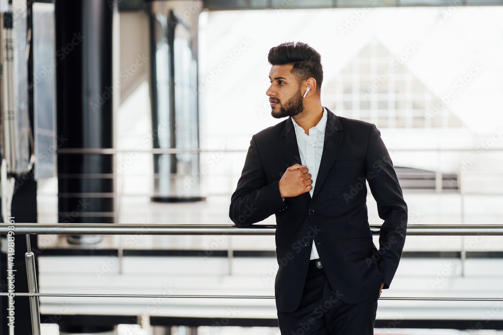 A modern Indian businessman in a modern office building, wearing a black suit, he speaks with wireless headphones