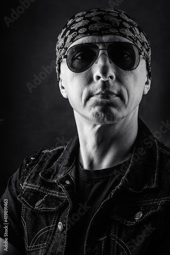 Unshaven, middle-aged man in a T-shirt, denim vest, bandana and sunglasses. Black and white portrait. Photo taken in studio