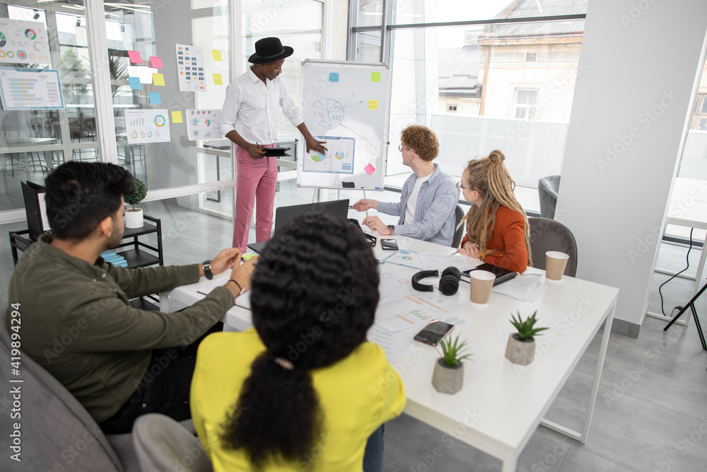 © sofiko14 - Young african man, standing near flipchart, and presenting details of joint project for multiracial team