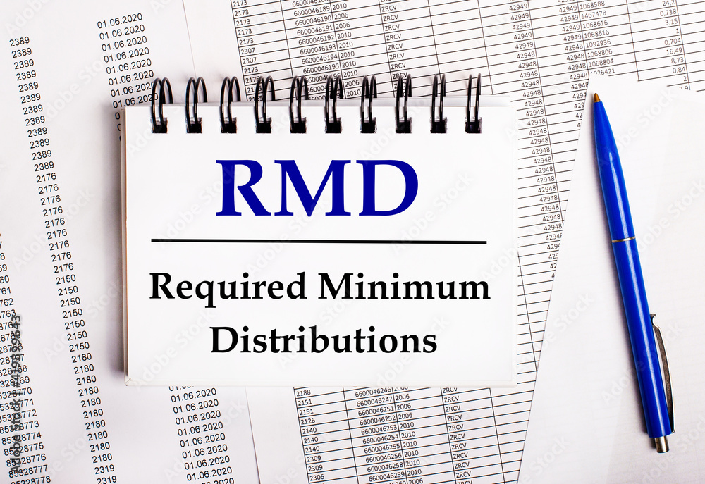 On the table are charts and reports, on which lie a blue pen and a notebook with the word RMD Required Minimum Distributions On the table are charts and reports, on which lie a blue pen and a notebook with the word RMD Required Minimum Distributions