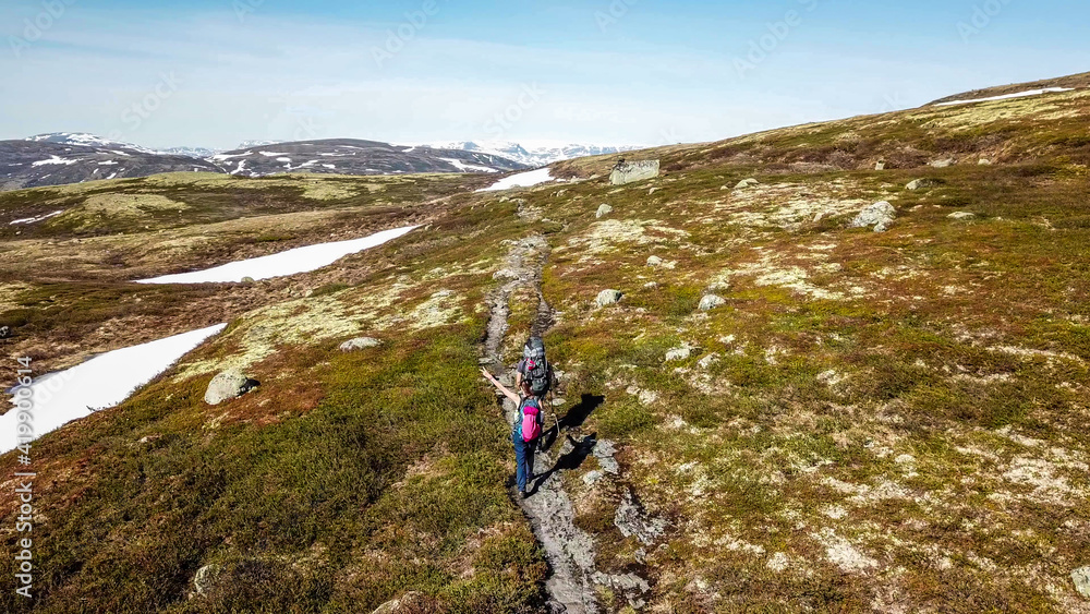 A couple hiking through the highlands of Eidfjors, Norway. Both of them carry big backpacks. Hike into the wilderness. Snow covers some parts of the slopes. Dry grass around the trail. Rough hike.
