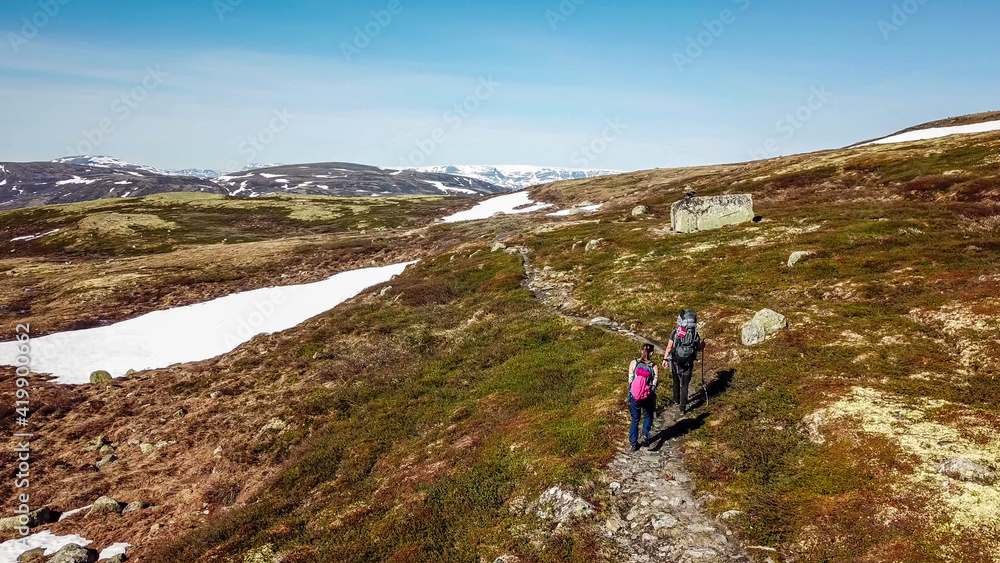 A couple hiking through the highlands of Eidfjors, Norway. Both of them carry big backpacks. Hike into the wilderness. Snow covers some parts of the slopes. Dry grass around the trail. Rough hike.