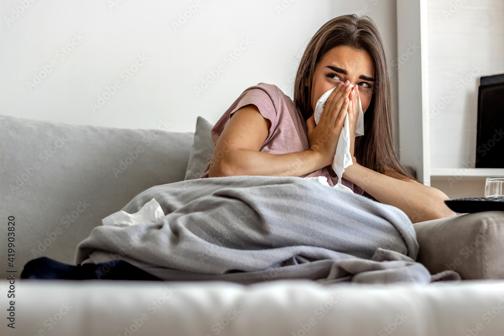 Shot of a frustrated woman using a tissue to sneeze in while being seated at home. Photo of sneezing woman in paper tissue. Attractive young girl feeling ill and blowing her nose with a tissue at home Shot of a frustrated woman using a tissue to sneeze in while being seated at home. Photo of sneezing woman in paper tissue. Attractive young girl feeling ill and blowing her nose with a tissue at home