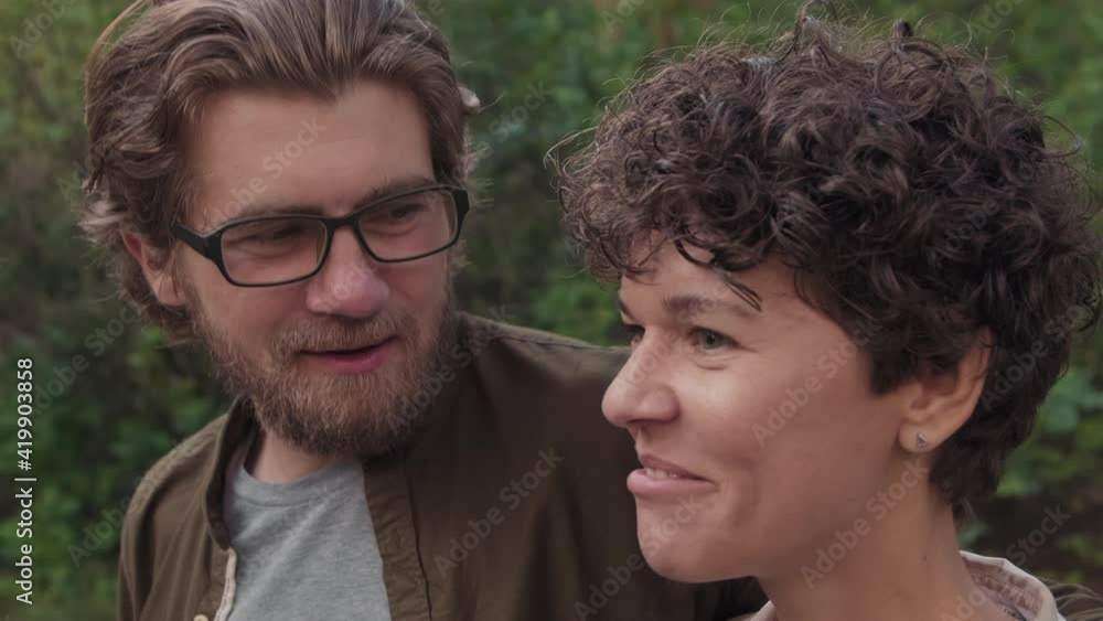 Handheld closeup of cheerful young caucasian couple smiling and talking while walking in picturesque green forest in summer enjoying nature Handheld closeup of cheerful young caucasian couple smiling and talking while walking in picturesque green forest in summer enjoying nature
