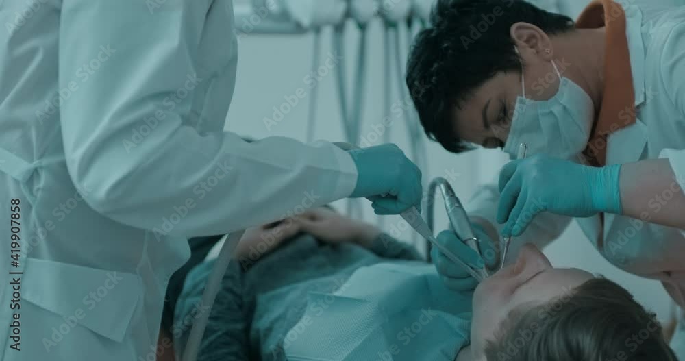 A female dentist in disposable mask is treating a male patient's teeth that lying in the dental chair using dental mirror and dental drill while the nurse is holding a saliva ejector. A female dentist in disposable mask is treating a male patient's teeth that lying in the dental chair using dental mirror and dental drill while the nurse is holding a saliva ejector.