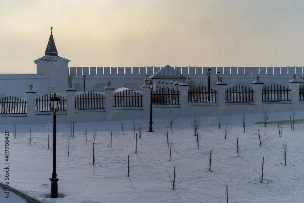 Young planting of fruit trees on the territory of the Tobolsk Kremlin (Siberia, Russia) on a cold winter evening. The battlements of the ancient kremlin, towers and lanterns in the distance 