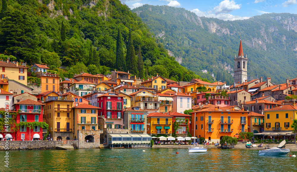 Varenna, Italy. Picturesque town at lake Como. Colourful motley Mediterranean houses at stone beach coastline among green trees. Popular health resort and touristic location. Summer day landscape.