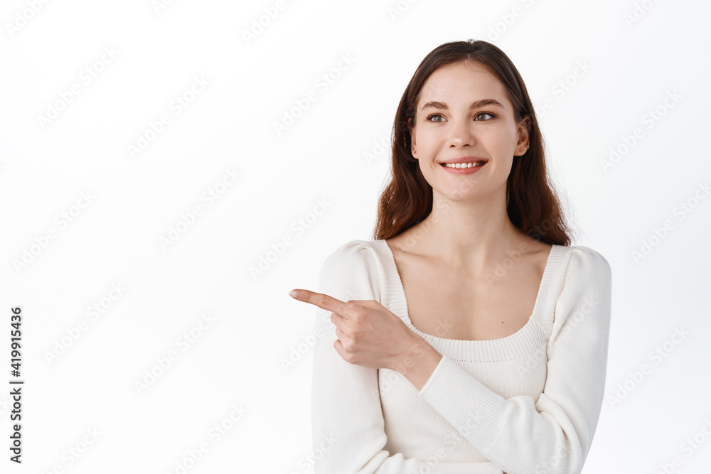 Portrait of young girl student smiling and looking hopeful at logo banner, pointing index finger aside, showing way, demonstrate banner, standing over white background Portrait of young girl student smiling and looking hopeful at logo banner, pointing index finger aside, showing way, demonstrate banner, standing over white background