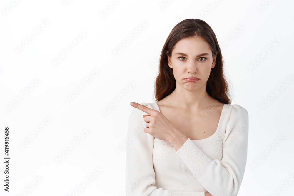 Blame, accusation. Displeased young woman pointing index finger aside, showing bad promo, sulking upset, looking disappointed, standing against white background Blame, accusation. Displeased young woman pointing index finger aside, showing bad promo, sulking upset, looking disappointed, standing against white background