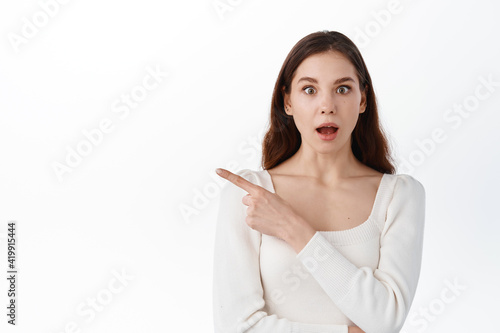 Surprised young woman look in awe, pointing index finger aside, showing awesome advertisement, demonstrate banner, wright your text here, standing against white background