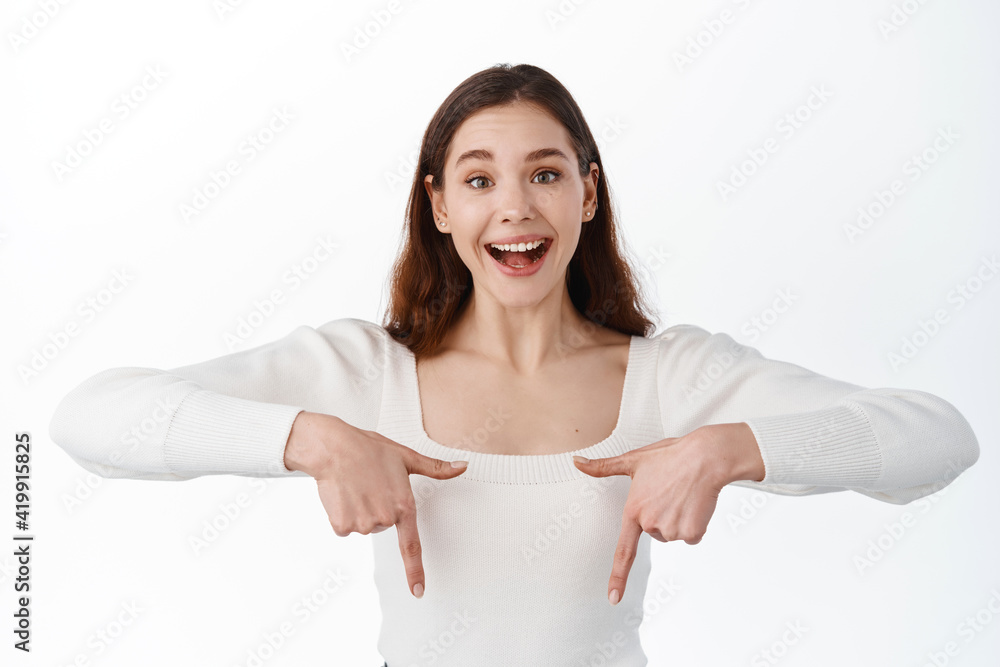 Image of happy young woman with hopeful face pointing fingers down, showing advertisement banner on copy space aside, smiling at camera, standing over white background Image of happy young woman with hopeful face pointing fingers down, showing advertisement banner on copy space aside, smiling at camera, standing over white background